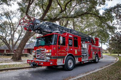 A red aerial fire truck with gray ladder sitting on a curved road with a green tree overhead.