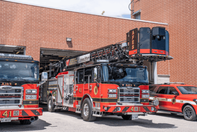 A red and black Pierce fire truck with a black aerial ladder parked on the pavement, exiting a fire station.