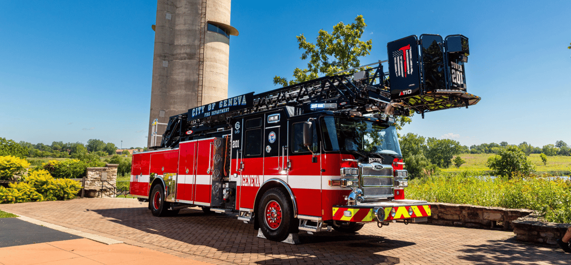 A red rear mount aerial fire truck with white stripe, sitting on a brick road with a silo and blue sky in the background.