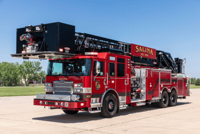A red Pierce fire truck with a black rear mounted aerial ladder sitting on concrete with blue sky and green trees in the background.