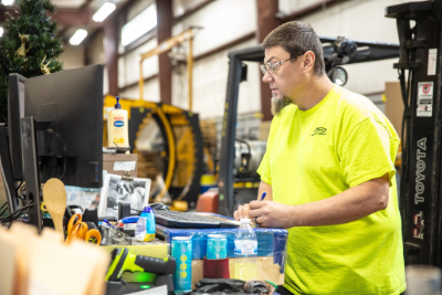 A man in a neon yellow shirt works at a computer screen in a factory setting.