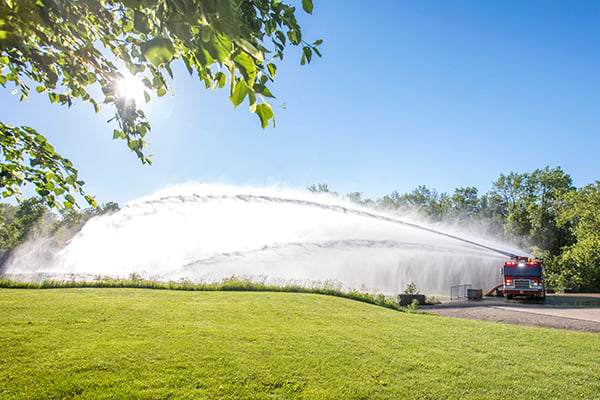 Pierce High Flow Industrial Apparatus Pumper Fire Truck spraying water from water pumps outside on a sunny day near grass. 