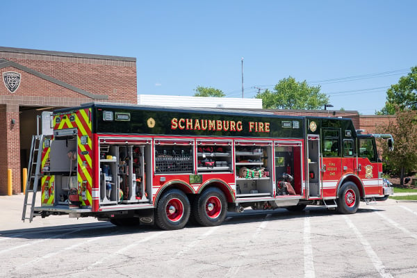 Passenger’s side of a Pierce Non-Walk-In Heavy-Duty Rescue Fire Truck parked outside in front of fire station with side and rear compartments open showing storage space. 