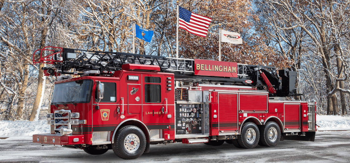 A red ladder fire truck with a black stripe and the work Bellingham sitting on snowy ground with bare trees and three flagpoles in the background.