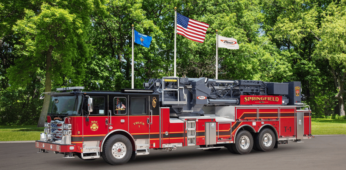 A red and black mid-mount fire truck with green trees and three flag poles in the background.