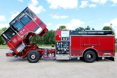 A red and black pumper fire truck is parked with the cab lifted, while also showing the driver side of the apparatus with a pump panel and compartments. 