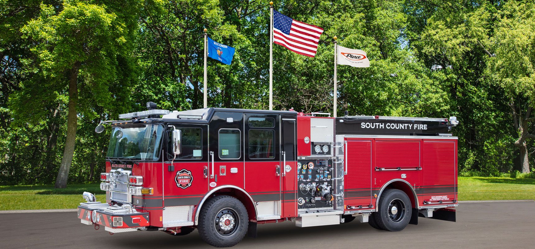 The red South County Fire pumper fire truck is parked on black asphalt with green trees and three flags in the background. 