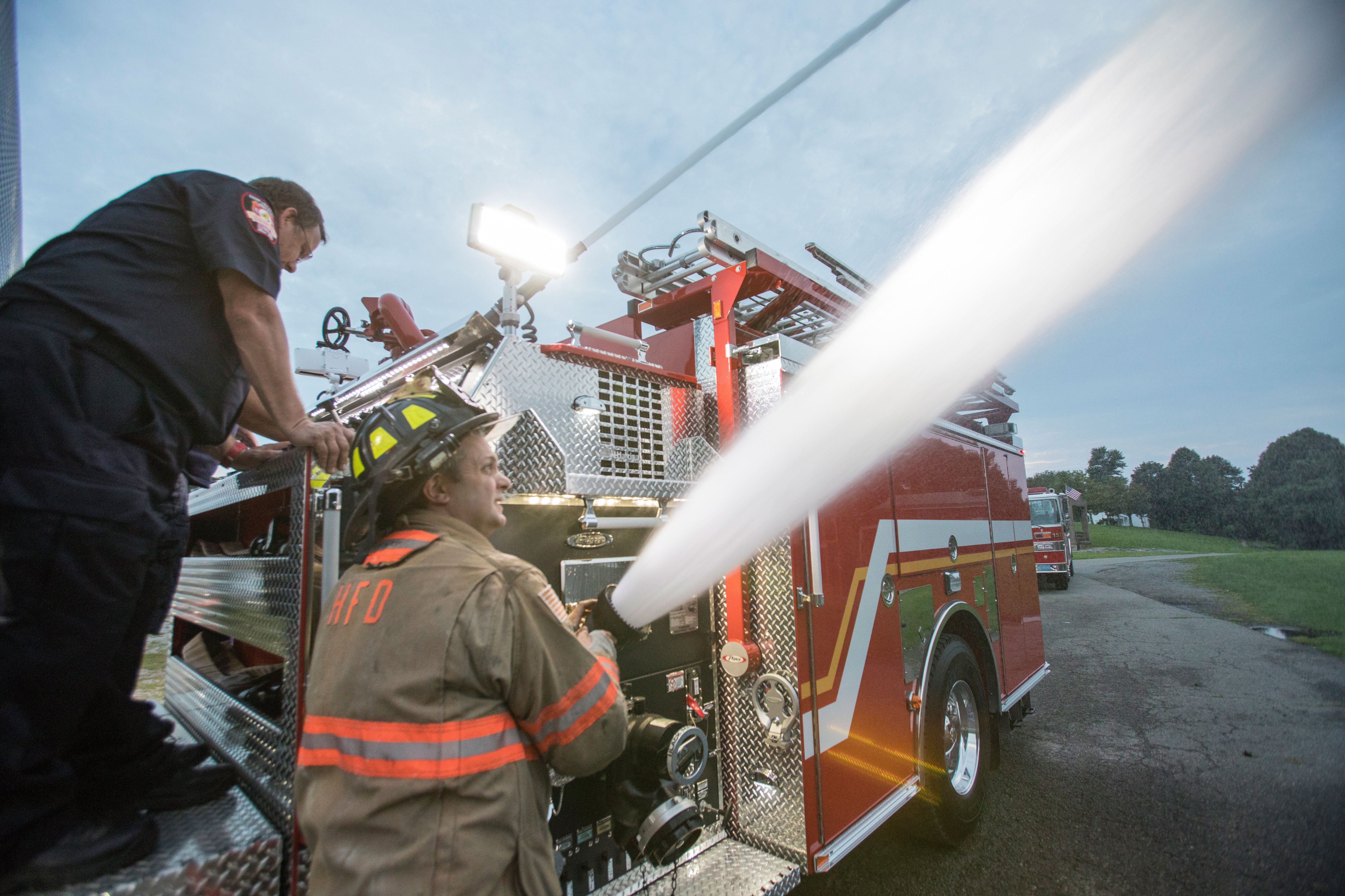 Two Firefighters pumping water from a top mounted pump panel on a Pierce Pumper Fire Truck parked outside.  