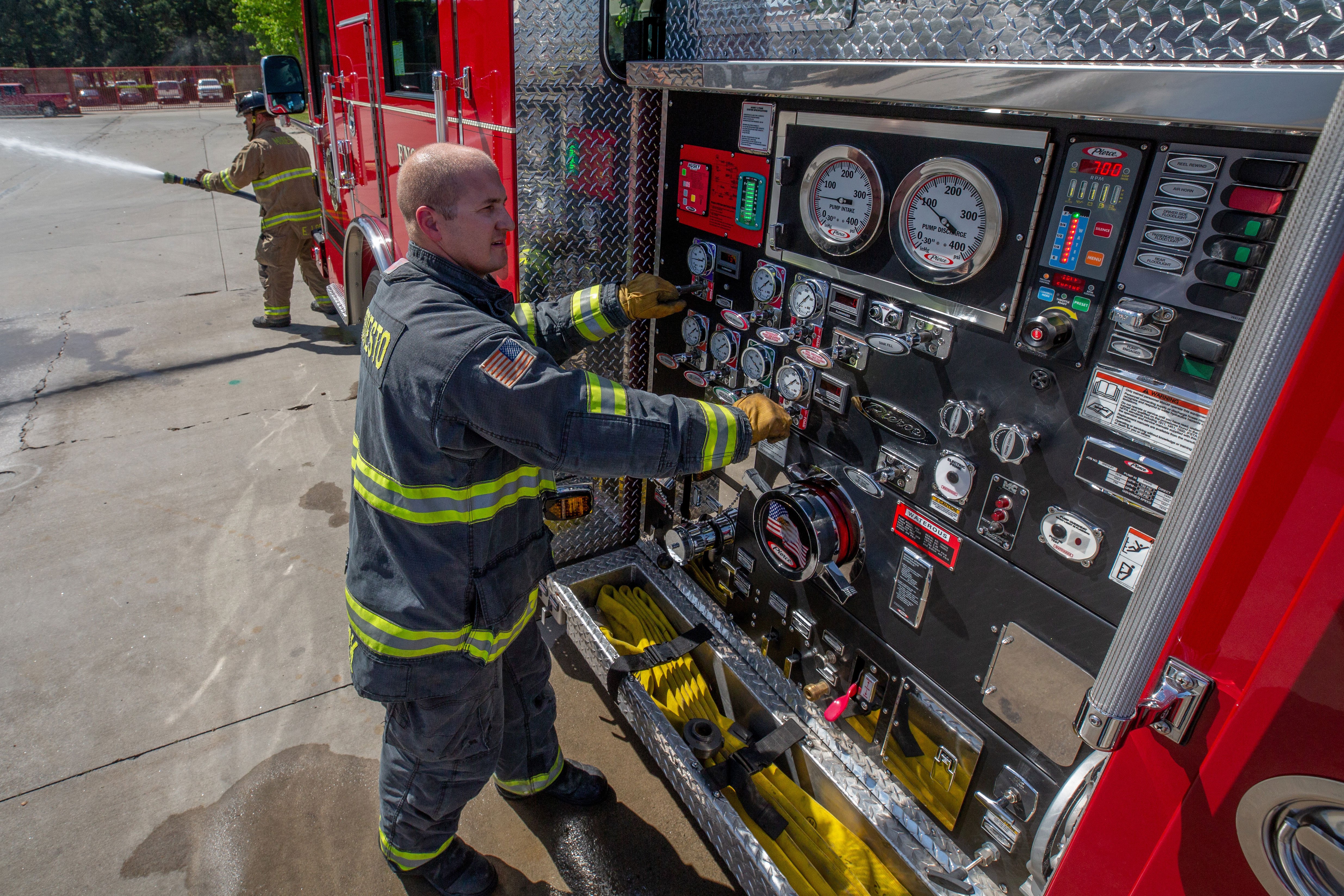 Two Firefighters spraying water and controlling the pump panel of a Pierce Pumper Fire Truck. 