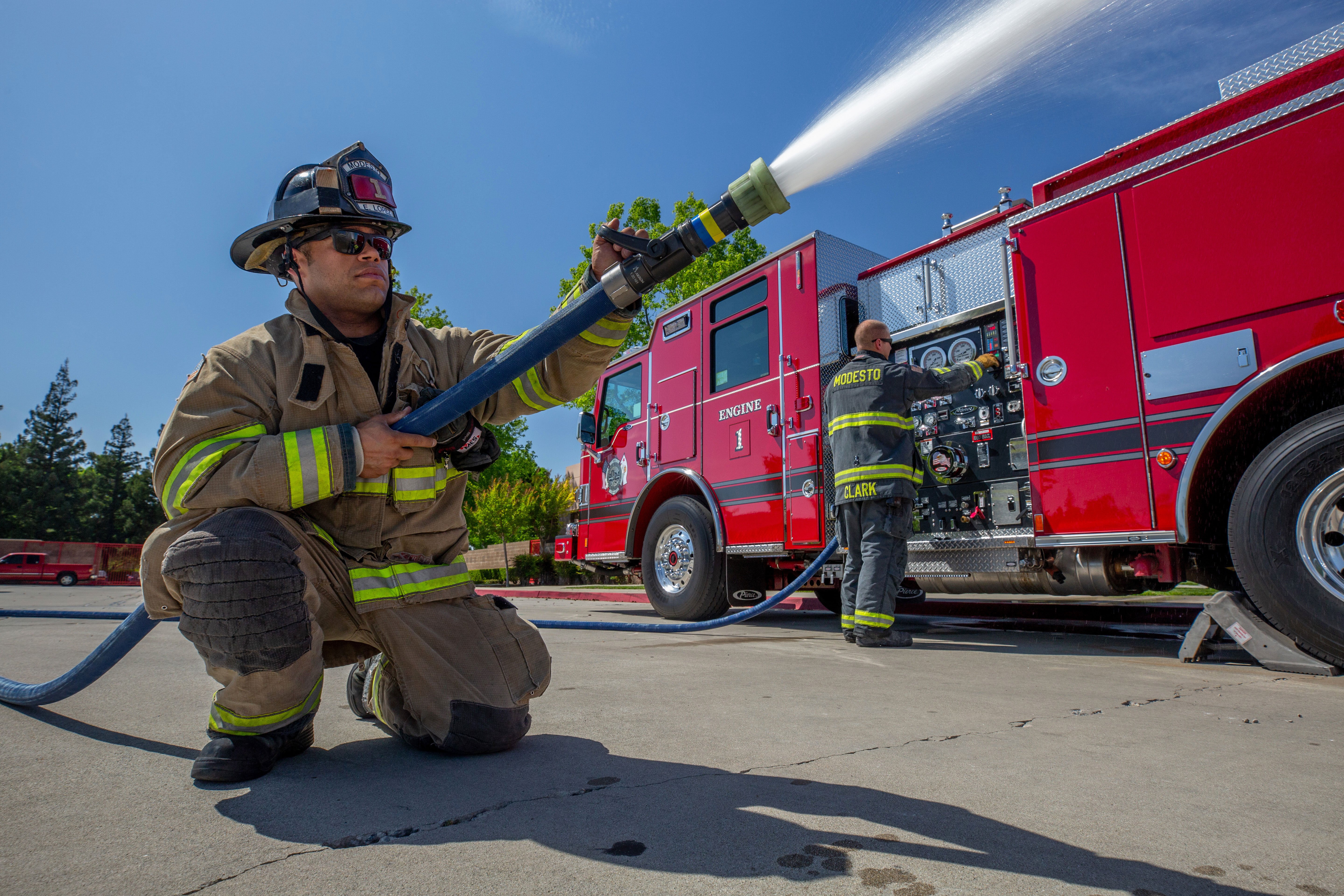 Firefighter outside with a hose pumping water out of a Pierce Pumper Fire Truck on a sunny day. 