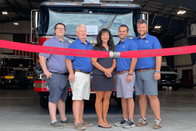 A team of people stand in front of a fire truck with a red fire hose reading "Reliant Fire" in front of them.