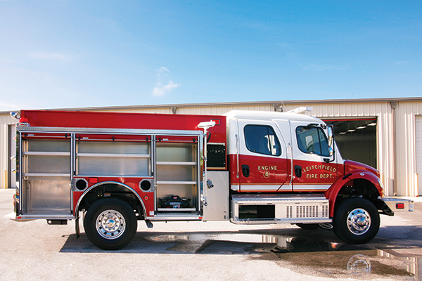 Passenger’s side of a Pierce Pumper Responder Fire Truck parked outside near a building with side compartments open showing storage space. 