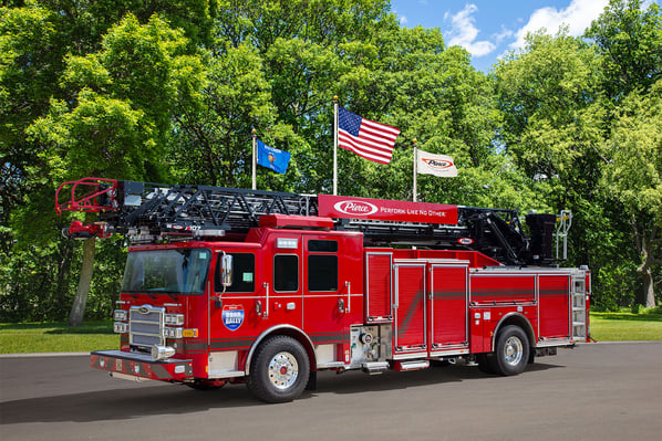 The front driver's side of an aerial fire truck in front of three flagpoles and trees on a sunny day.