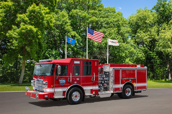 The front driver's side of a fire truck parked in front of three flagpoles and trees on a sunny day.