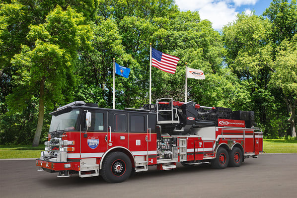 The front driver's side of an aerial fire truck in front of three flagpoles and trees on a sunny day.