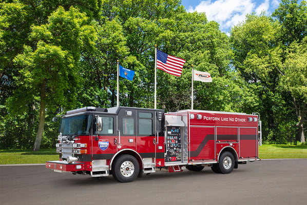 The front driver's side of a fire truck parked in front of three flagpoles and trees on a sunny day.