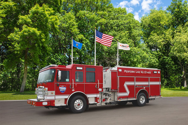 The front driver's side of a fire truck parked in front of three flagpoles and trees on a sunny day.