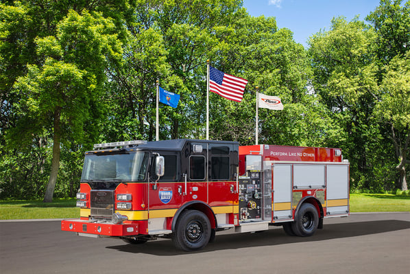 The front driver's side of a fire truck parked in front of three flagpoles and trees on a sunny day.