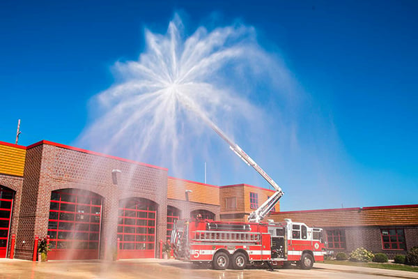 Pierce Fire Truck parked outside on a sunny day in front of a Fire Station with the High-Reach Snozzle extended out in front of the truck spraying water into the air. 