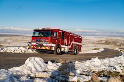 The front drivers side of a fire truck driving on a winding road in the winter with snow on the ground and mountains behind.
