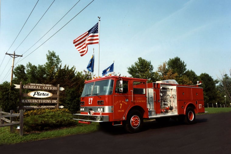 Pierce Manufacturing Arrow fire apparatus parked outside beside the Pierce sign and the flag poles.