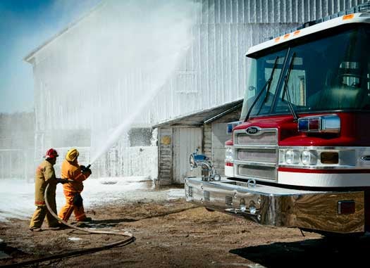 Two firefighters spraying water on a building from a hose beside a Pierce Manufacturing firetruck. 