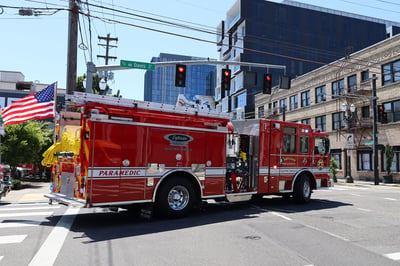 The officer's side of a Volterra electric fire truck with an American flag on the rear, driving through an intersection in a city to an emergency.