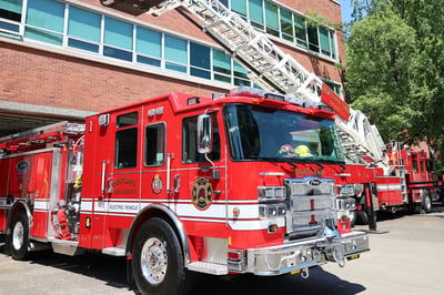 The front officer's side of a Volterra electric fire truck parked in front of a bring fire station next to an aerial fire truck.