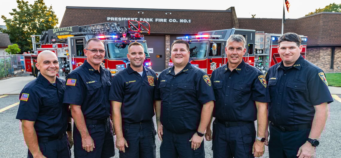 Six firefighters in blue uniforms stand smiling with two fire trucks and the President Park Fire Co. No.1 building in the background.