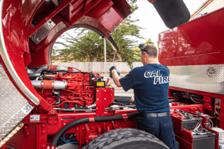 A Cal Fire firefighter in a blue shirt is standing in front of an open fire truck hood, inspecting the red engine with his hands. The truck is parked outdoors.