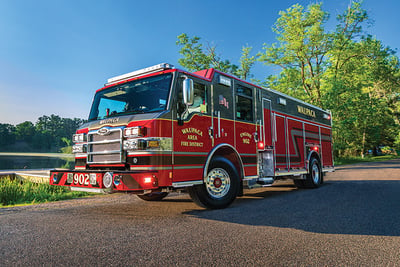 A red fire truck is driving on an asphalt road next to a lake with green trees in the background.