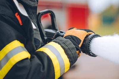 A firefighter in black turnout gear with yellow stripes holds a nozzle spraying firefighter foam.