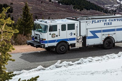 A drone view of a white fire truck as it drives through snow-capped  mountains and evergreen trees.