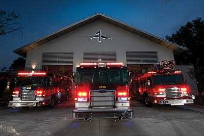 Three fire trucks are parked in front of a fire station with emergency lights on at night.