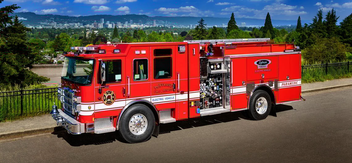 Portland Fire & Rescue’s red Pierce Volterra electric fire truck is parked on a road overlooking trees, the city and mountains in the background.