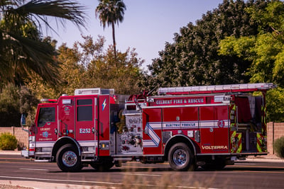 A red electric fire truck initiates a left turn on an urban street with palm trees and greenery in the background.   