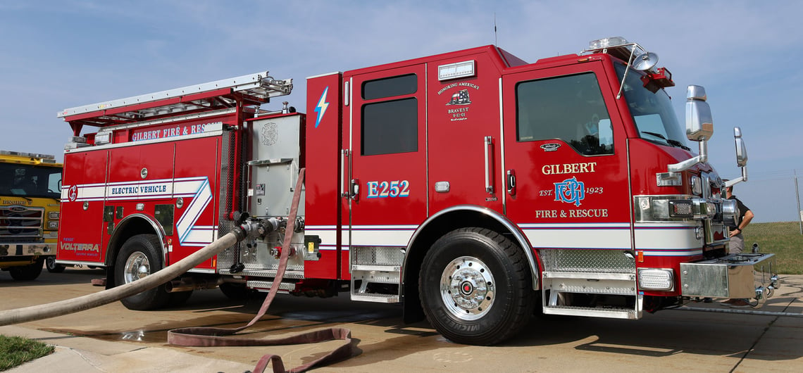 A red electric pumper truck with hoses attached and a blue sky in the background.