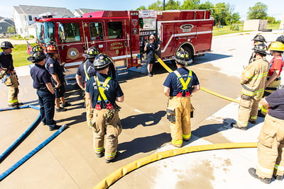 A group of firefighters in station gear stand outside in front of a red electric fire truck as one firefighter leads a training session. 
