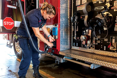 A female firefighter plugs an electric fire truck into the overhead power source in a fire station.