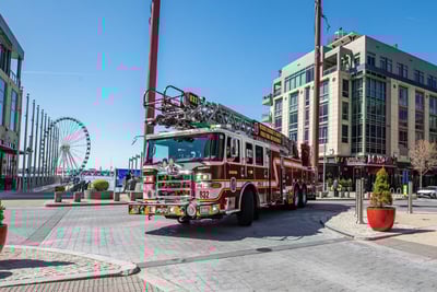 A red and white aerial fire truck is parked in a commercial area with buildings and a Ferris wheel in the background. 