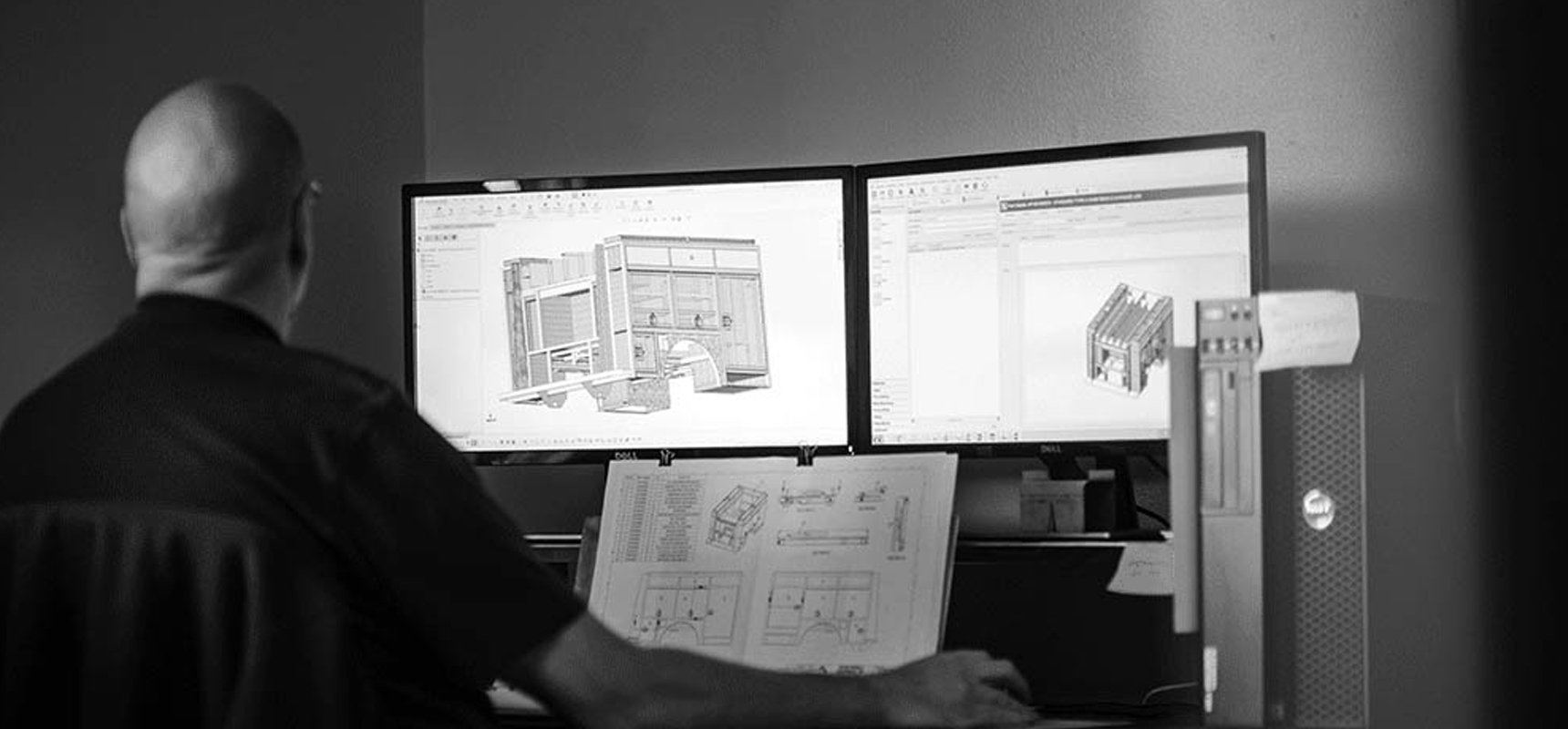 A black and white photo shows a man in a black shirt sitting at a desk looking at two screens with wildland fire truck renderings and a paper document with additional schematics. 