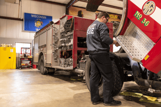A firefighter is inspecting the side of a red and silver fire truck inside a garage bay. A flag that reads "WISCONSIN 1848" hangs on the wall behind the truck, near a yellow storage cabinet.