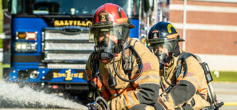 Two firefighters holding a hose spraying water with a blue fire apparatus in the background