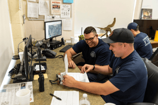 Two firefighters in blue shirts are sitting at a desk and reviewing documents and binders together. One firefighter is pointing to a page and smiling