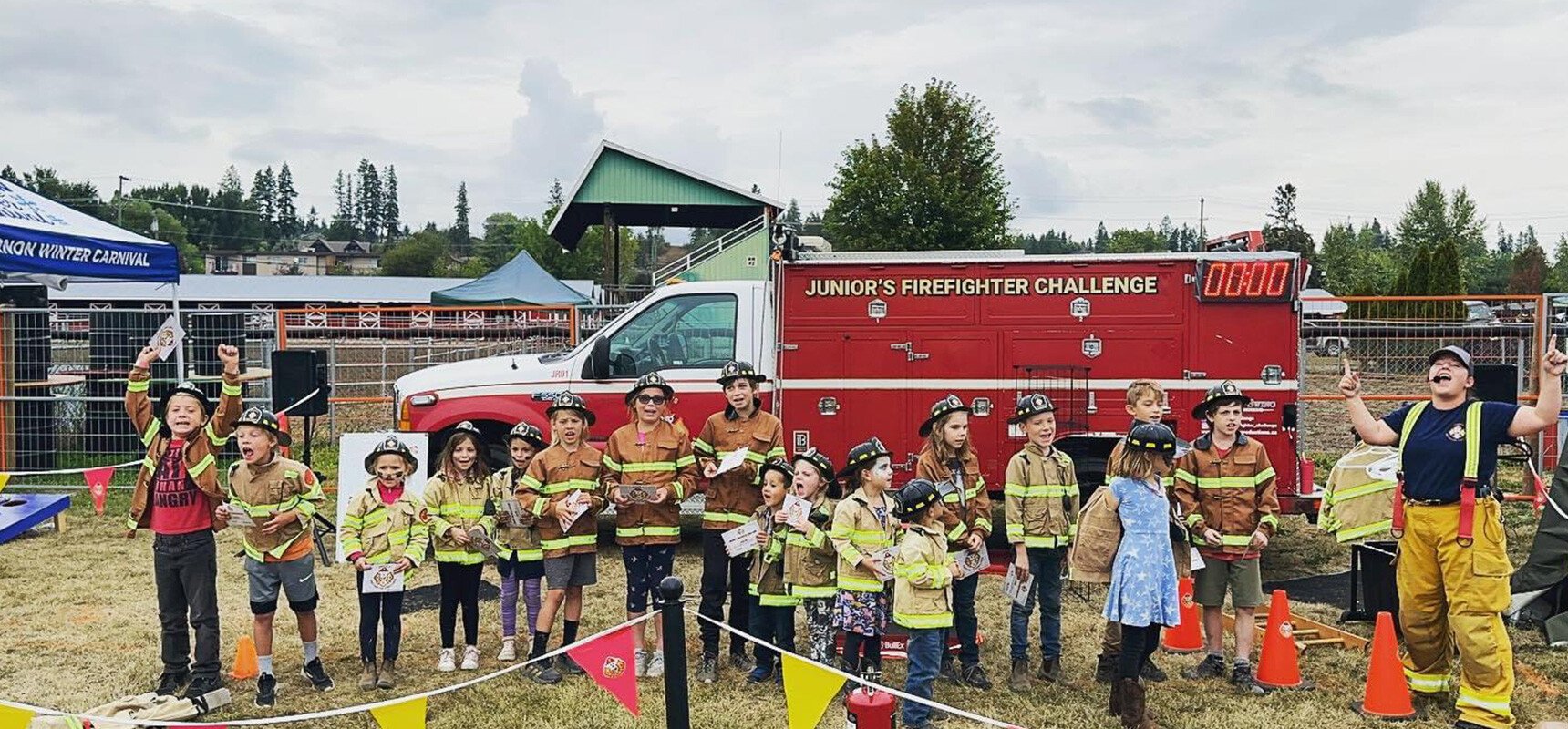 A group of kids in firefighter jackets stand in the grass in front of a red ambulance.