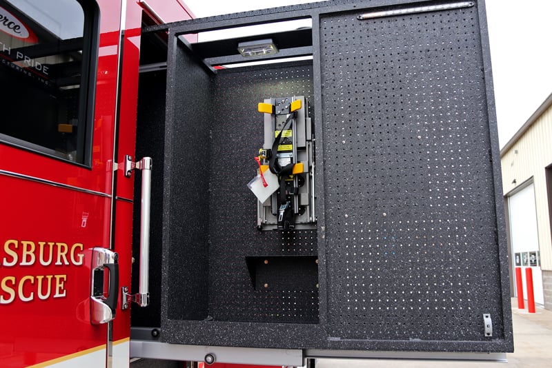 A red fire truck is parked and a slide-out isolated compartment is pulled out behind the operator’s door showing a peg board with tools and storage. 