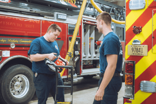 Two firefighters are standing in a fire station garage bay, with a large red and gray fire truck behind them. One firefighter is holding a piece of rescue equipment and the other is looking on.