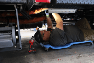 A mechanic, holding a drill in one hand, is lying on the ground and working on the undercarriage of a fire truck.