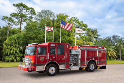 A red fire truck with black stripe sitting on the pavement with green trees and a blue sky, with three flag poles in the background.