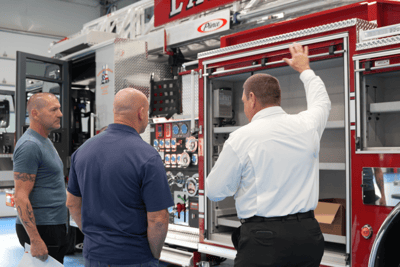 A Ten-8 Fire &amp; Safety team member opens a fire truck compartment to show the store space to two firefighters.